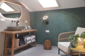 Une salle de bain avec un meuble-lavabo en bois, un grand miroir, un évier et un miroir rond au-dessus du meuble-lavabo. On y trouve un panier en osier sur le sol et une chaise en osier avec un coussin.