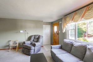 A living room with a grey sofa, a cushioned armchair, a wooden chair, a lamp, and a window with curtains.