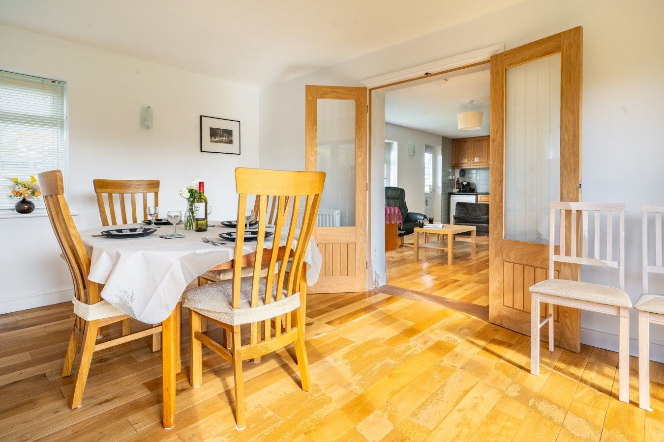 Un coin repas avec des chaises en bois et une table dressée pour deux personnes, une cuisine en arrière-plan et un couloir menant à une autre pièce.