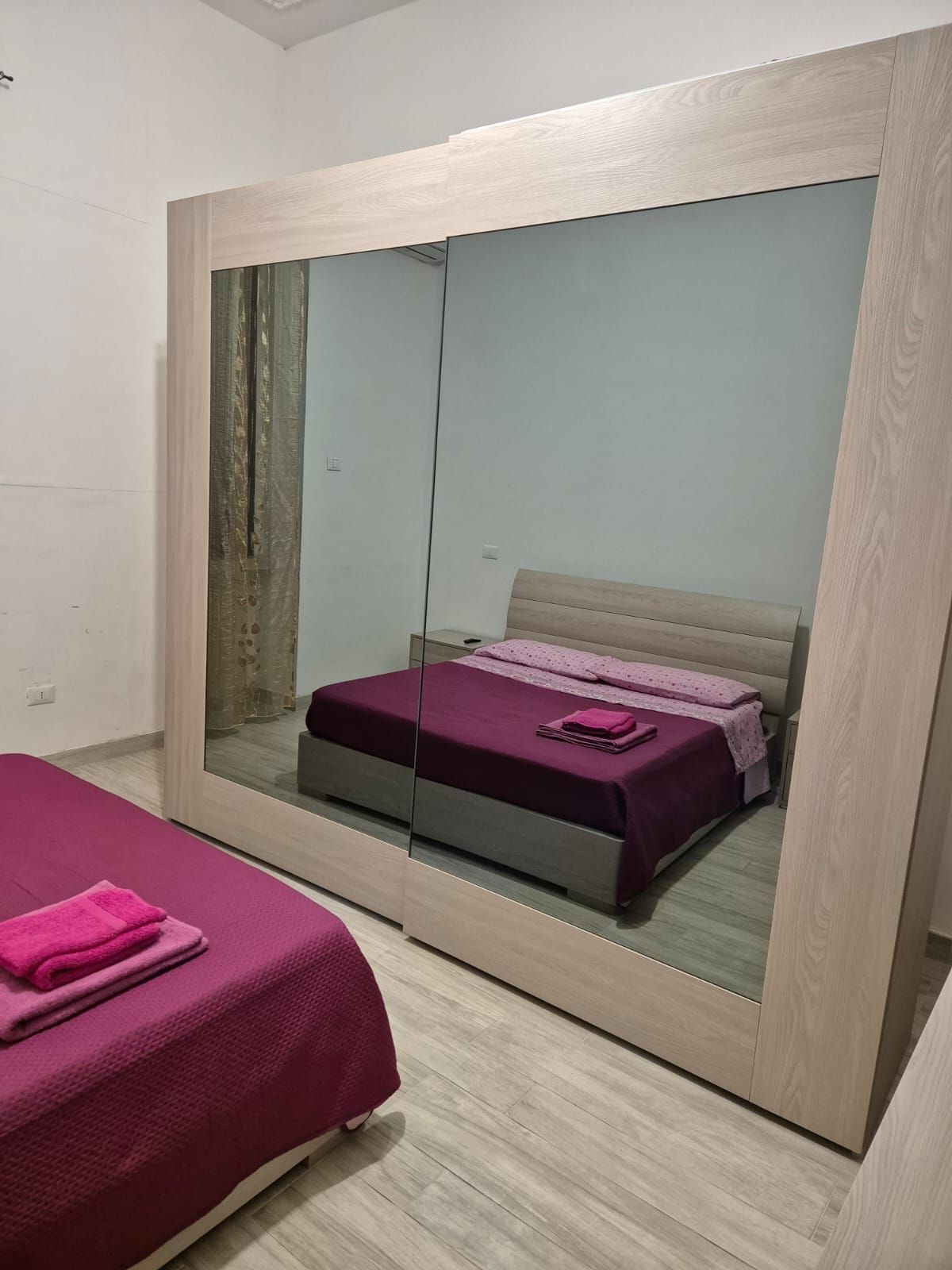 Une chambre avec un grand miroir, un lit recouvert d’une courtepointe rose et une armoire en bois.