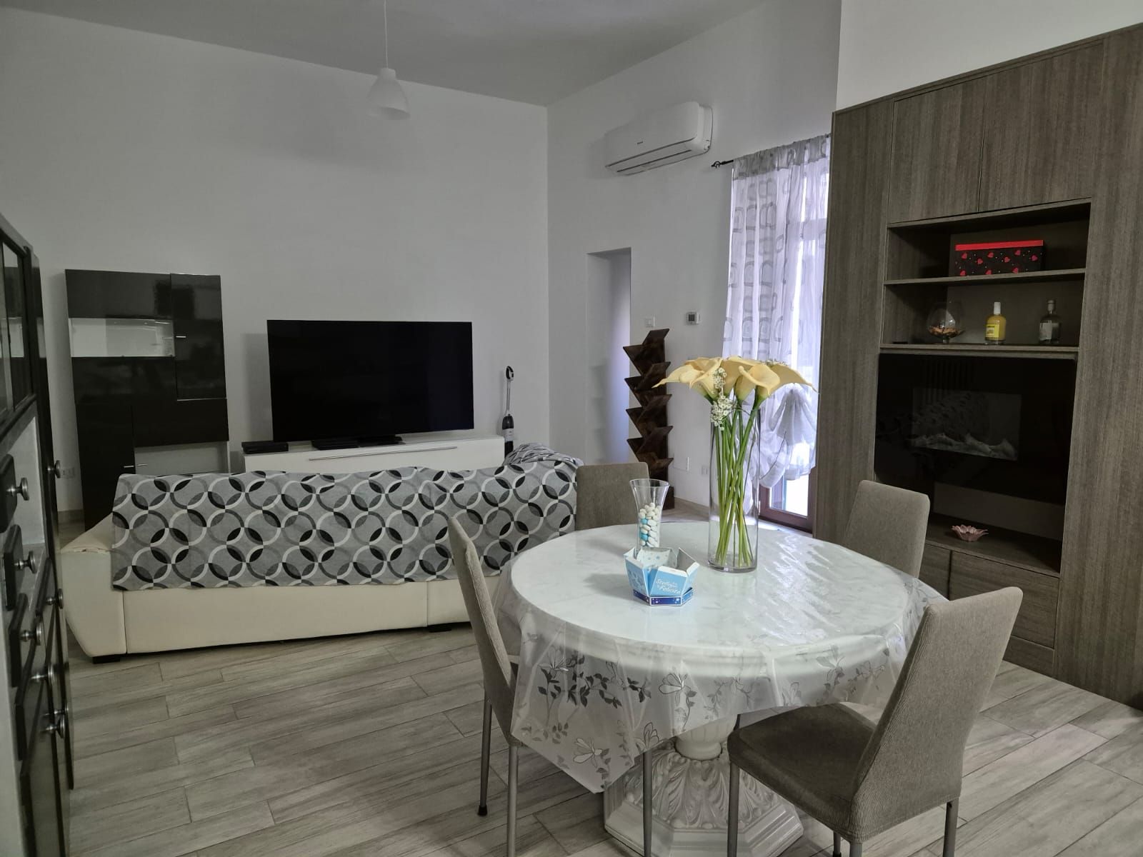Ein modernes Wohnzimmer mit einem Flachbildfernseher, einer Couch mit geometrischem Muster, einem Esstisch mit Stühlen und einer Vase mit Blumen.