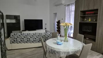 Ein modernes Wohnzimmer mit einem Flachbildfernseher, einer Couch mit geometrischem Muster, einem Esstisch mit Stühlen und einer Vase mit Blumen.