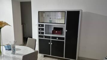 A room with a white table, chairs, a cabinet with drawers, a clock on the wall, and a bed with a patterned cover.