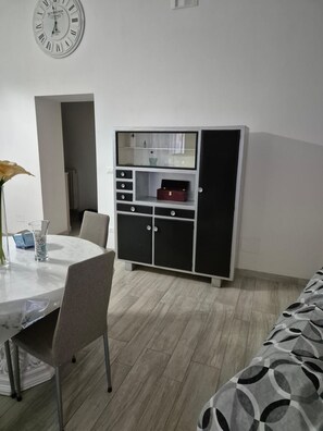 Ein Zimmer mit weißem Tisch, Stühlen, einer Kommode mit Schubladen, einer Uhr an der Wand und einem Bett mit gemustertem Überwurf.