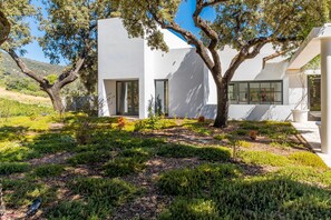 A modern building with a white exterior, large windows, and a well-maintained garden with various plants and a tree. - Cubo's Villa La Caldera / private pool / gym / vineyards / luxury experiences / free Wi-Fi (Ronda)