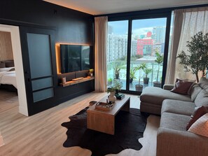 A modern living room with a flat-screen TV, a sofa, a coffee table, and a view of a cityscape through large windows. - Luxury 2BR/2BA Tampa Channelside Downtown - Gym + Sauna + Pool  (Tampa)