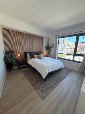 A bedroom with a large bed, a nightstand, a lamp, a potted plant, and a view of buildings through a window. - Luxury 2BR/2BA Tampa Channelside Downtown - Gym + Sauna + Pool  (Tampa)