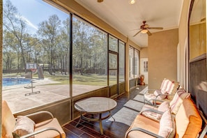 A spacious room with large windows, a ceiling fan, and a view of a backyard with a pool and trees. - Sleeps 16 Pool Slide Family Retreat (Porter)