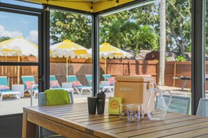 Una mesa de madera con una cafetera, una caja y vasos en un patio soleado con sombrillas y sillones de descanso.