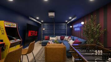 A room with a blue sofa, a foosball table, a large flat-screen TV, and a Pac-Man arcade machine.