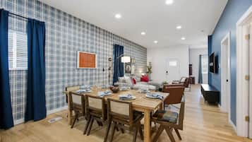 Un comedor con una mesa de madera preparada para cuatro personas, flanqueada por cortinas azules, un sofá y un televisor al fondo.