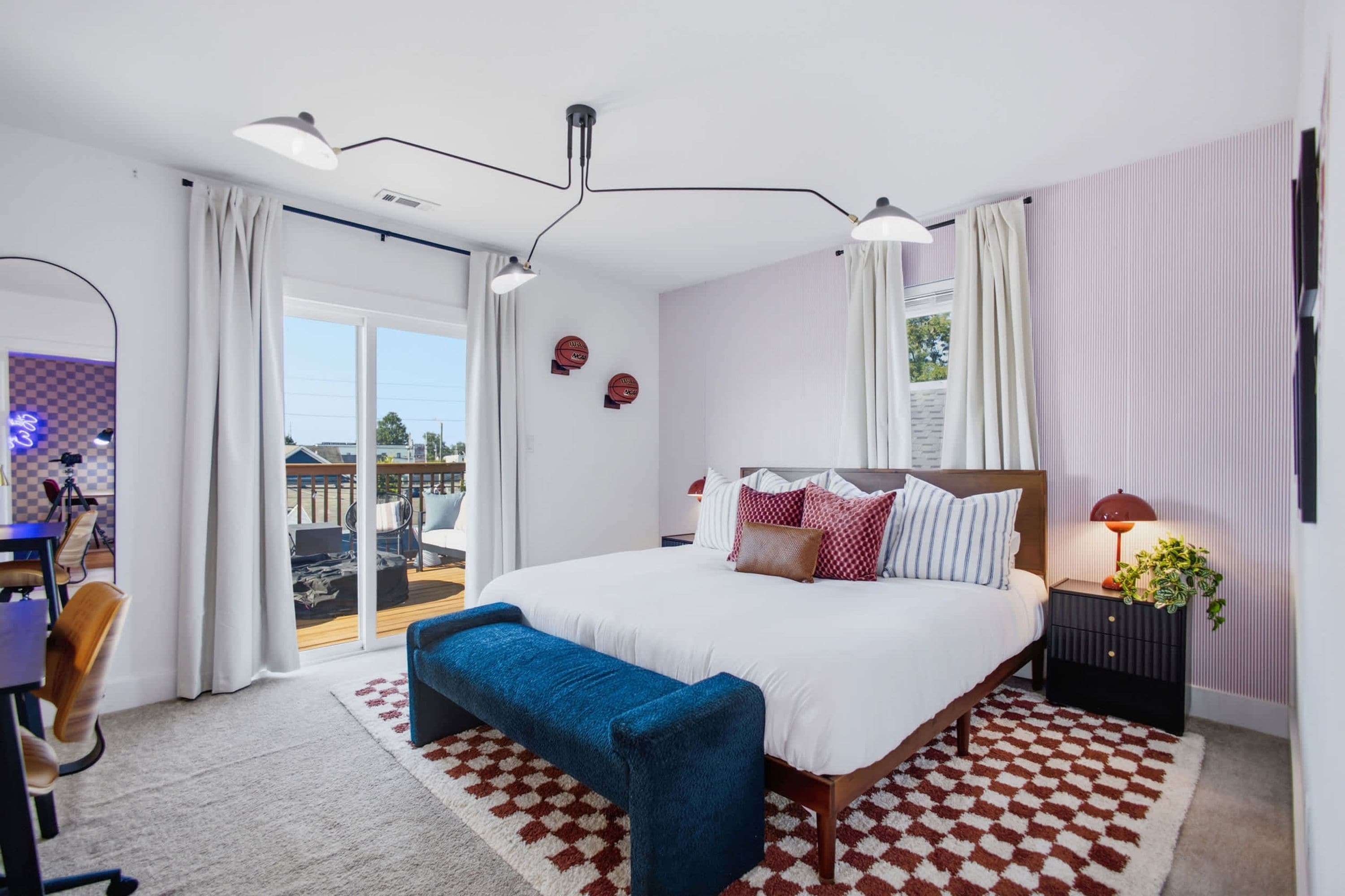 A bedroom with a large bed, a desk with a chair, a bench, a nightstand with a lamp, and a view of the outdoors.