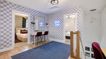 A hotel room with a checkered wall pattern, a bed, a desk with two chairs, a blue rug, and a staircase.