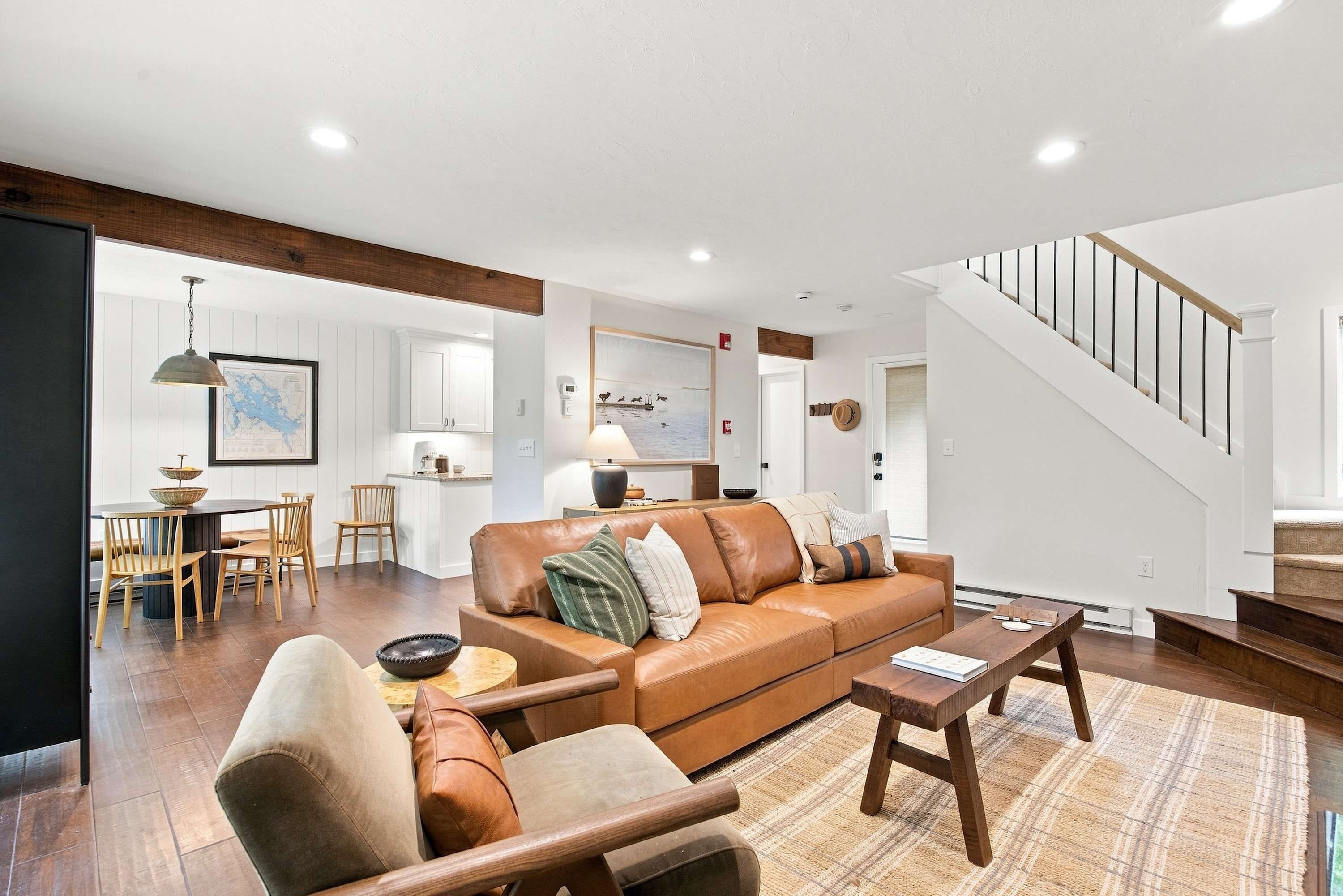 A modern living room with a brown leather sofa, wooden coffee table, and a dining area with wooden chairs and a table.