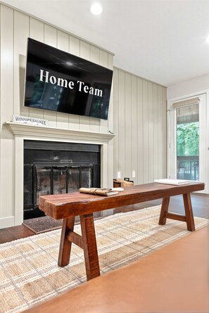 Ein Wohnzimmer mit Kamin, einem an der Wand befestigten Fernseher, der 'Home Team' zeigt, und einer hölzernen Bank.
