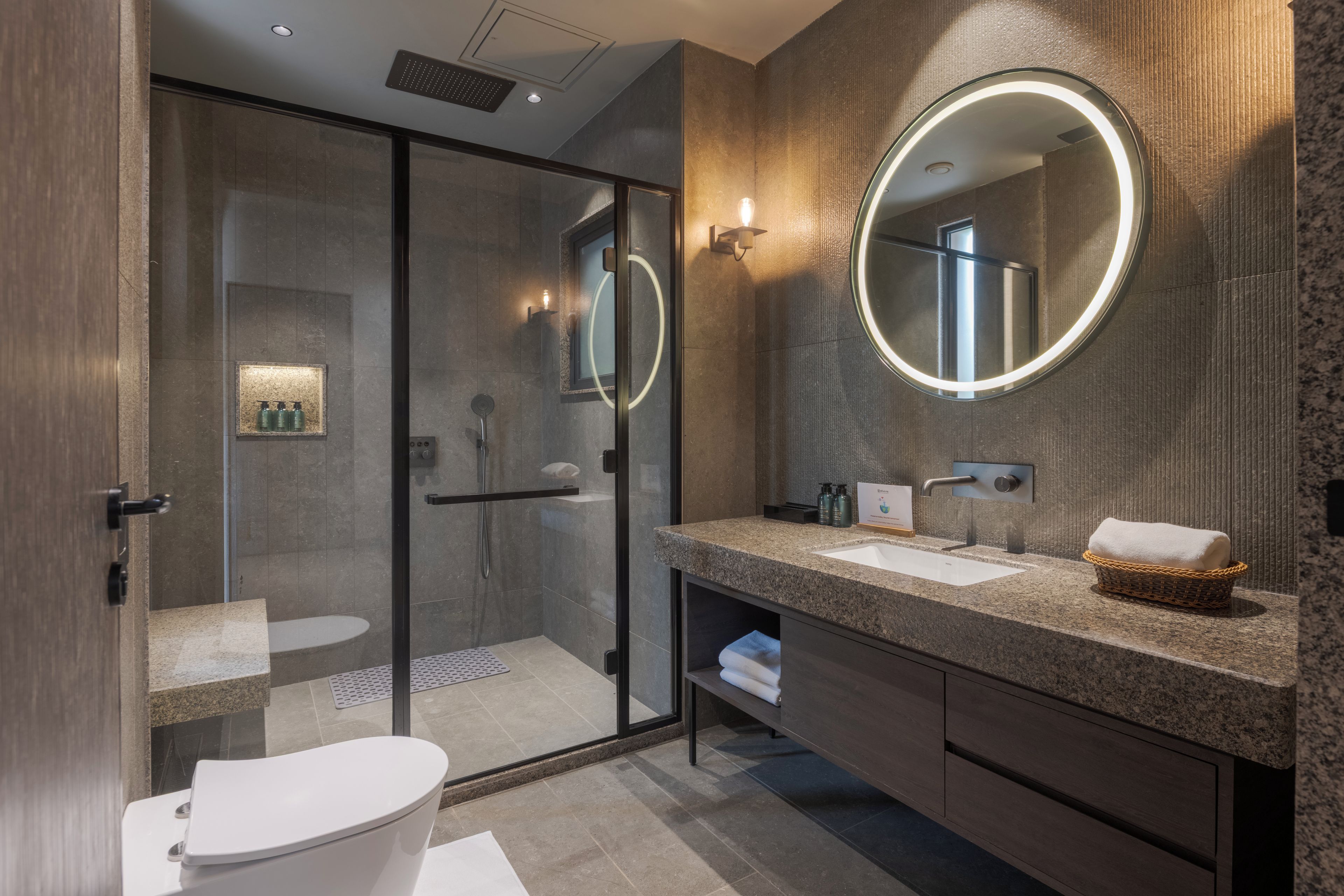 A modern bathroom with a glass-enclosed shower, a round mirror, and a granite countertop.