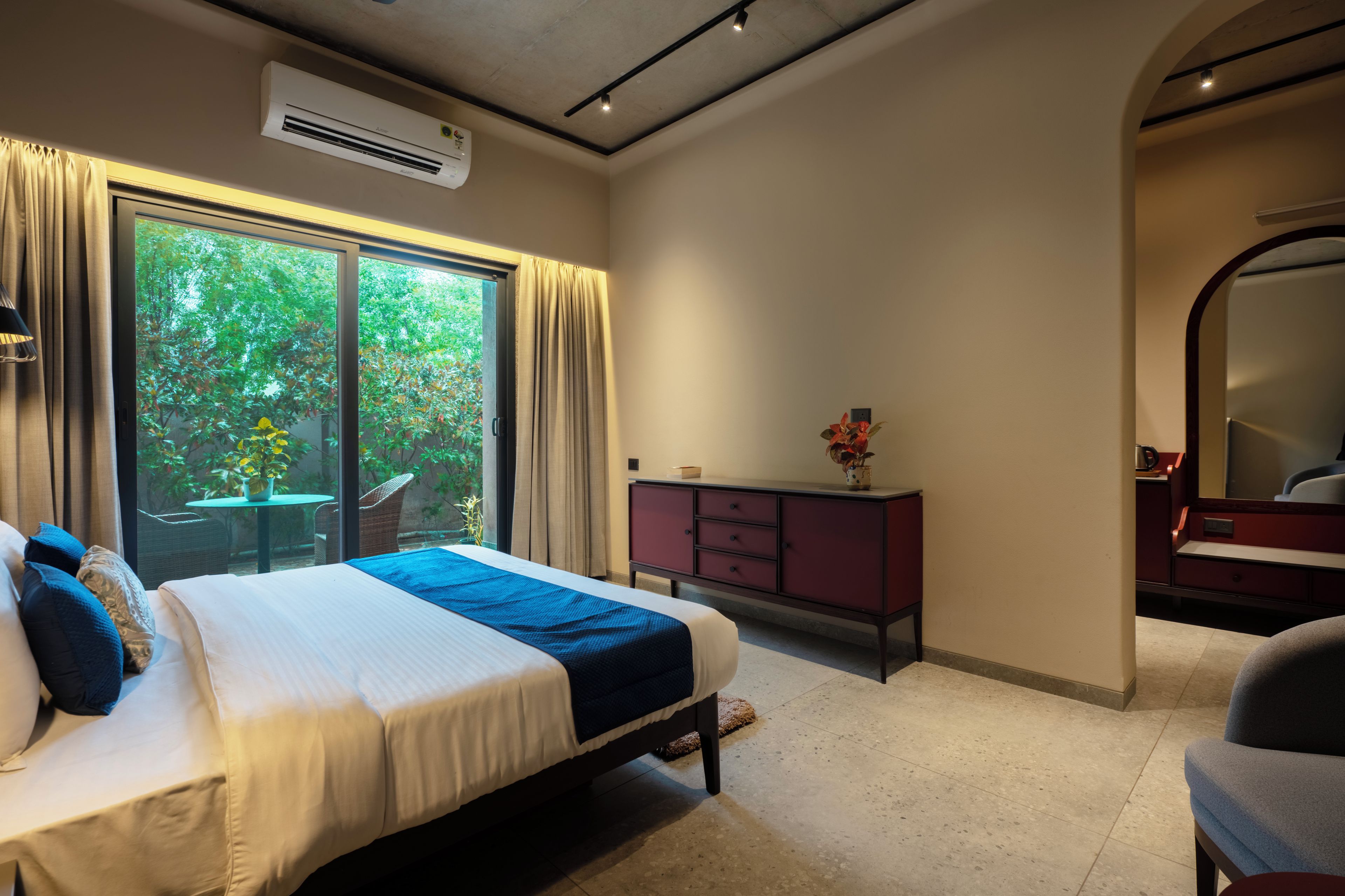 A hotel room with a bed, a dresser, a mirror, and a window with a view of greenery.