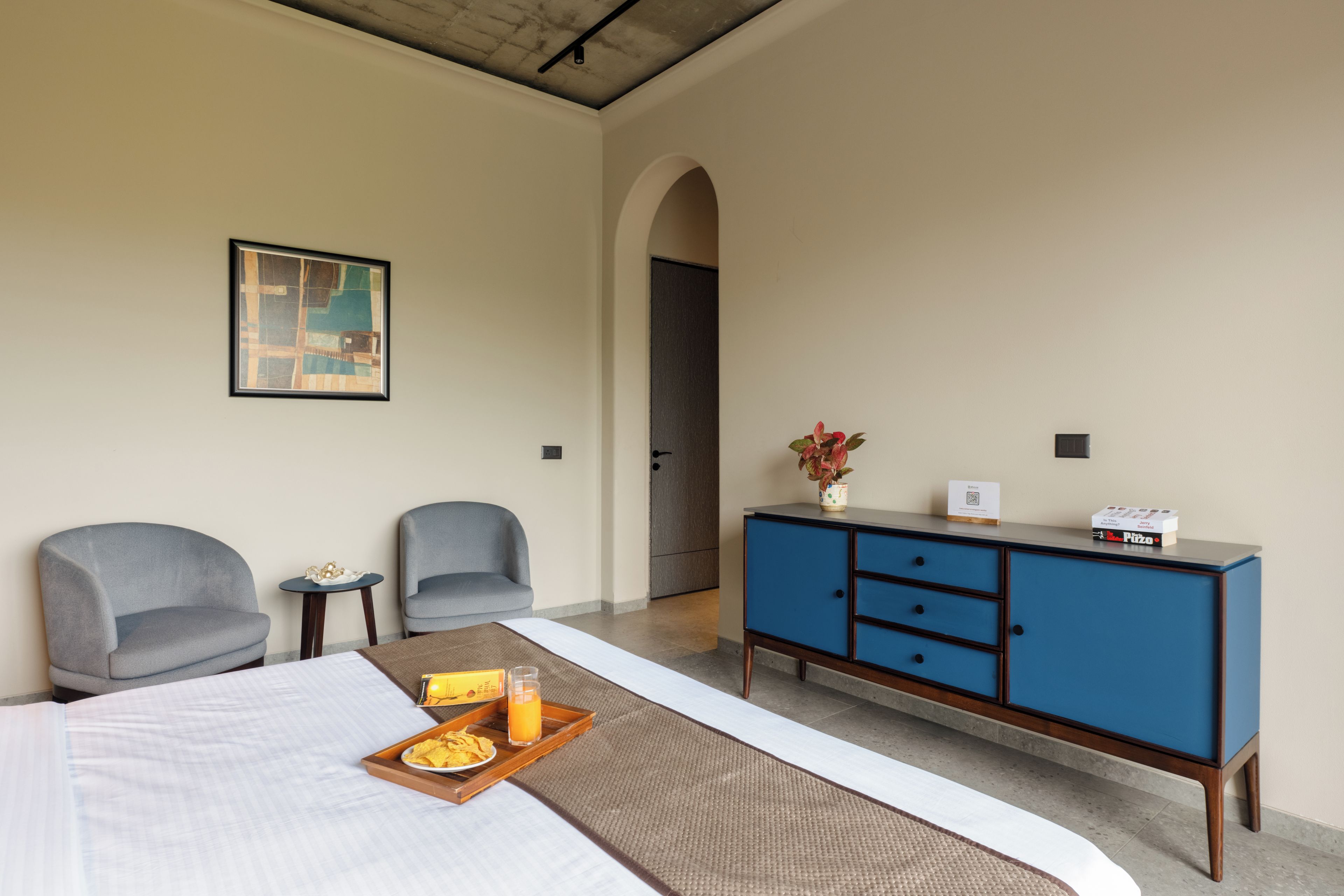 A hotel room with a bed, two armchairs, a small table with a tray of snacks, a blue cabinet, and a painting on the wall.