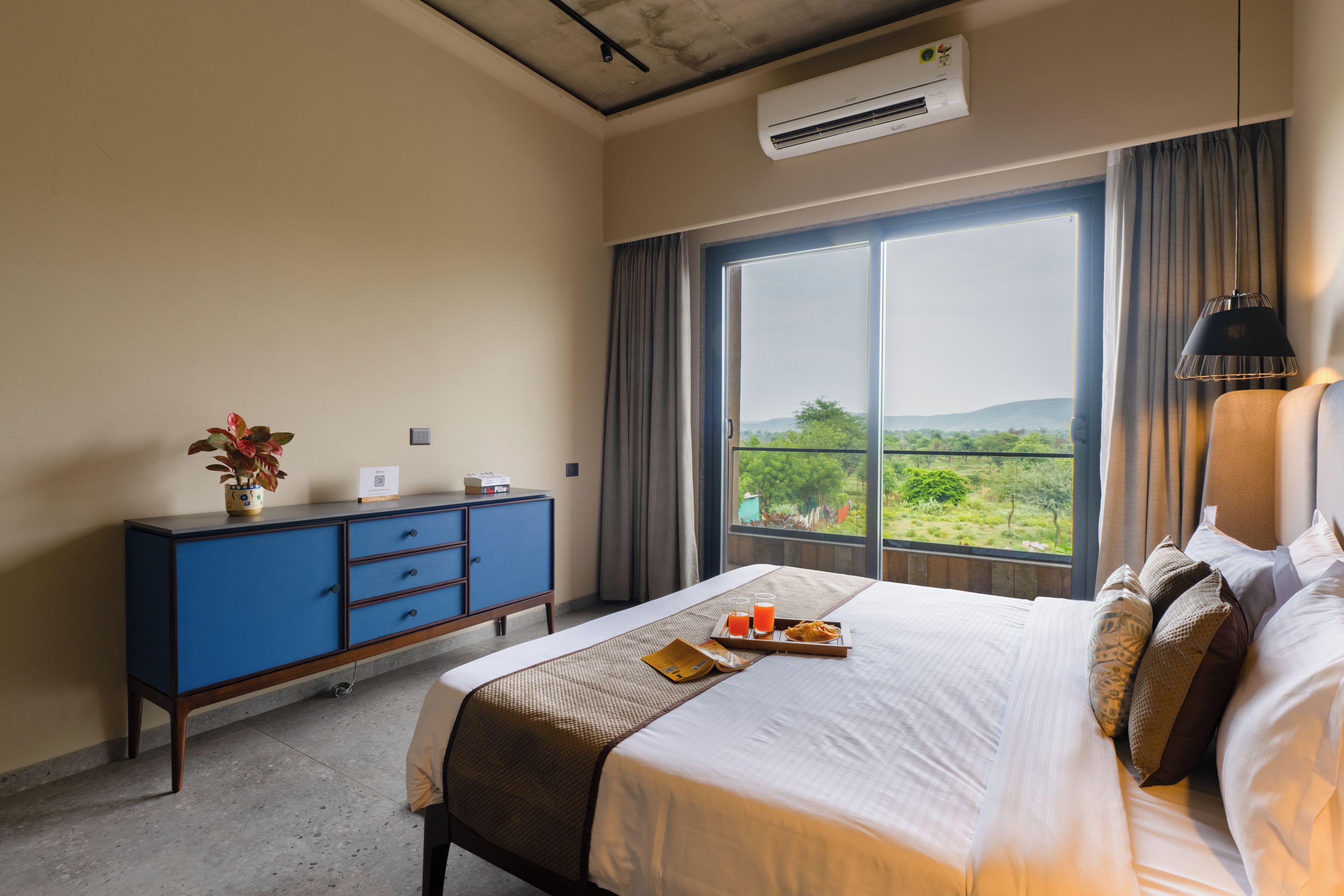 Quarto de hotel com uma cama grande, uma cômoda azul, uma janela com vista para vegetação e um aparelho de ar condicionado.