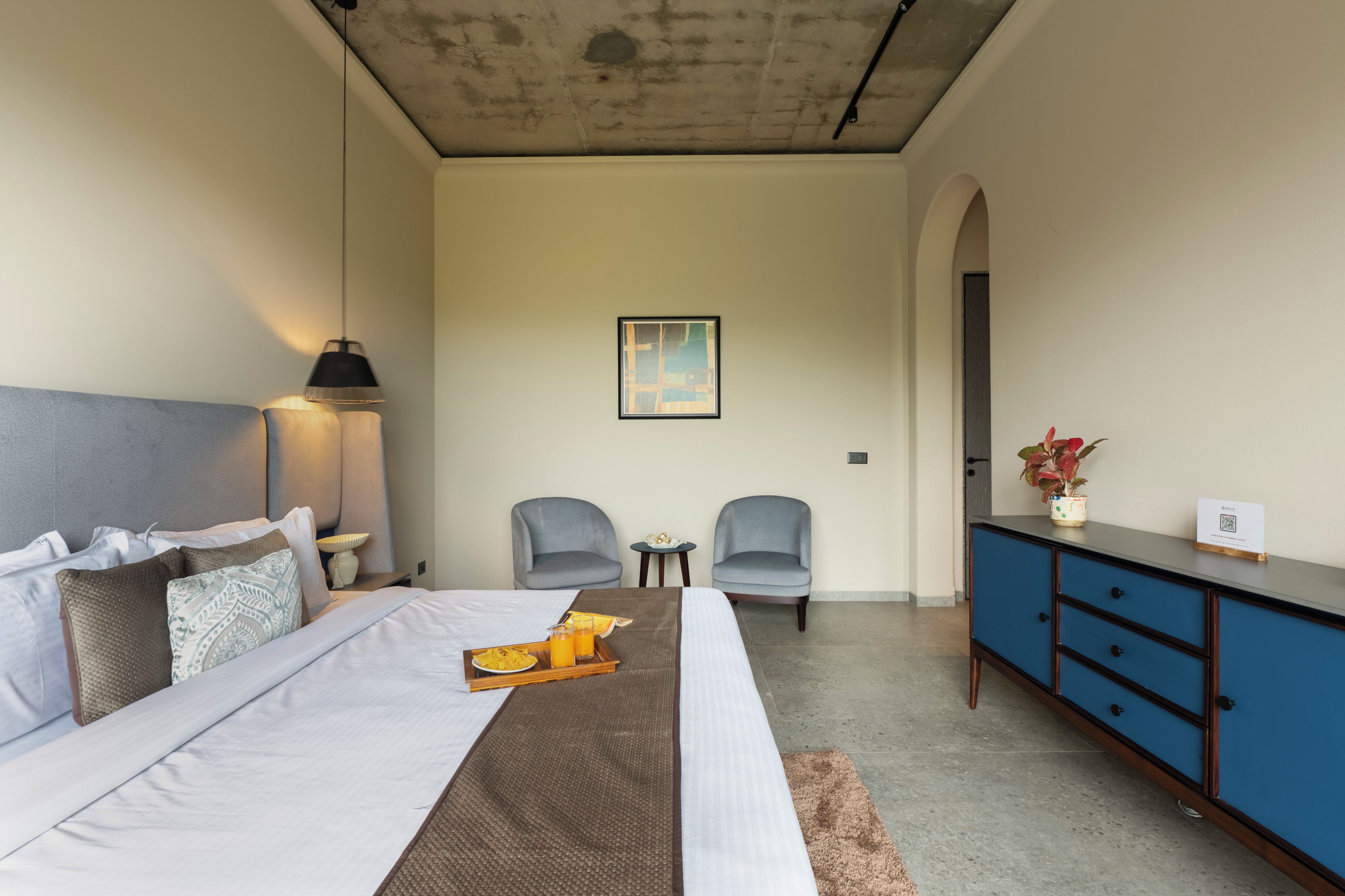 A hotel room with a bed, bedside table, lamp, two armchairs, a small table, a cabinet, and a painting on the wall.