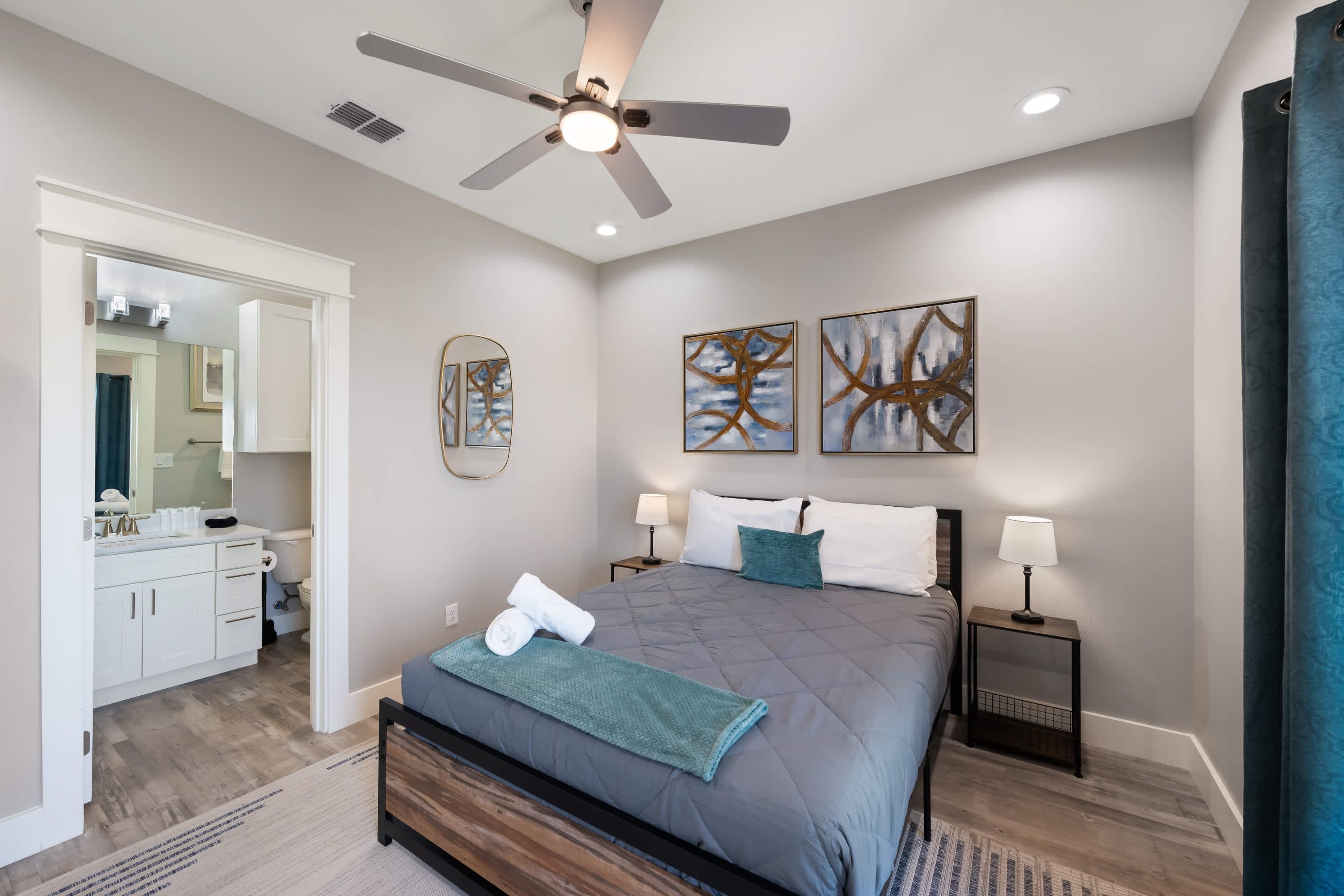Un dormitorio moderno con cama, mesitas de noche, ventilador de techo, obras de arte y un baño visible a través de una puerta abierta.