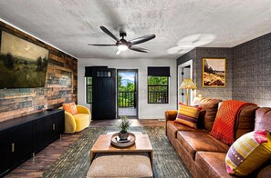 A modern living room with a ceiling fan, a large painting, a sofa, a coffee table, and a view of the outdoors through a door. - Rolling Hills Retreat Hot Tub Fire Pit Sauna (Sevierville)