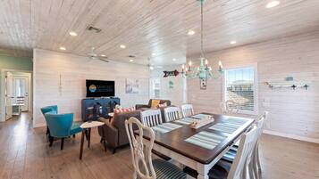A spacious room with a wooden ceiling, a dining table with chairs, a television, and a chandelier.