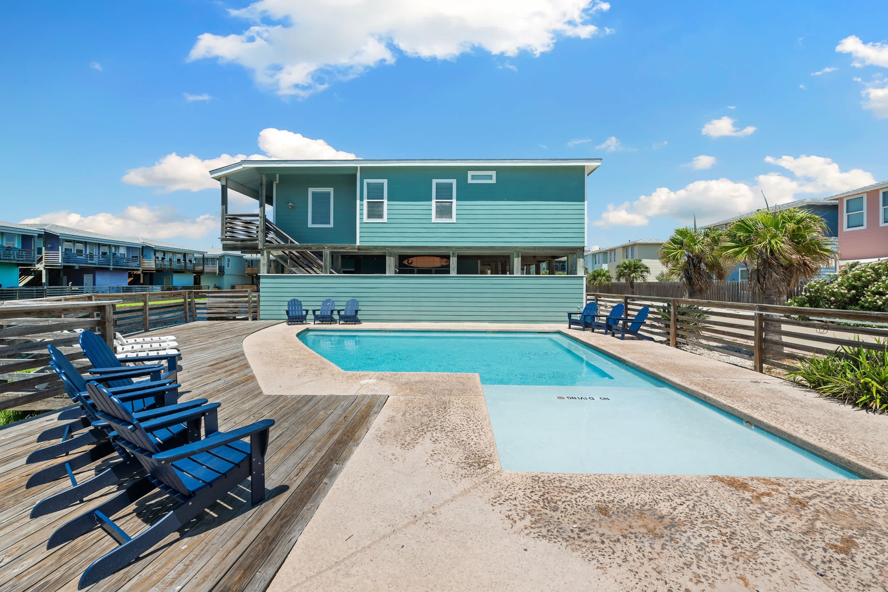 A pool area with a wooden deck, lounge chairs, and a building with a staircase.