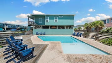 A pool area with a wooden deck, lounge chairs, and a building with a staircase.