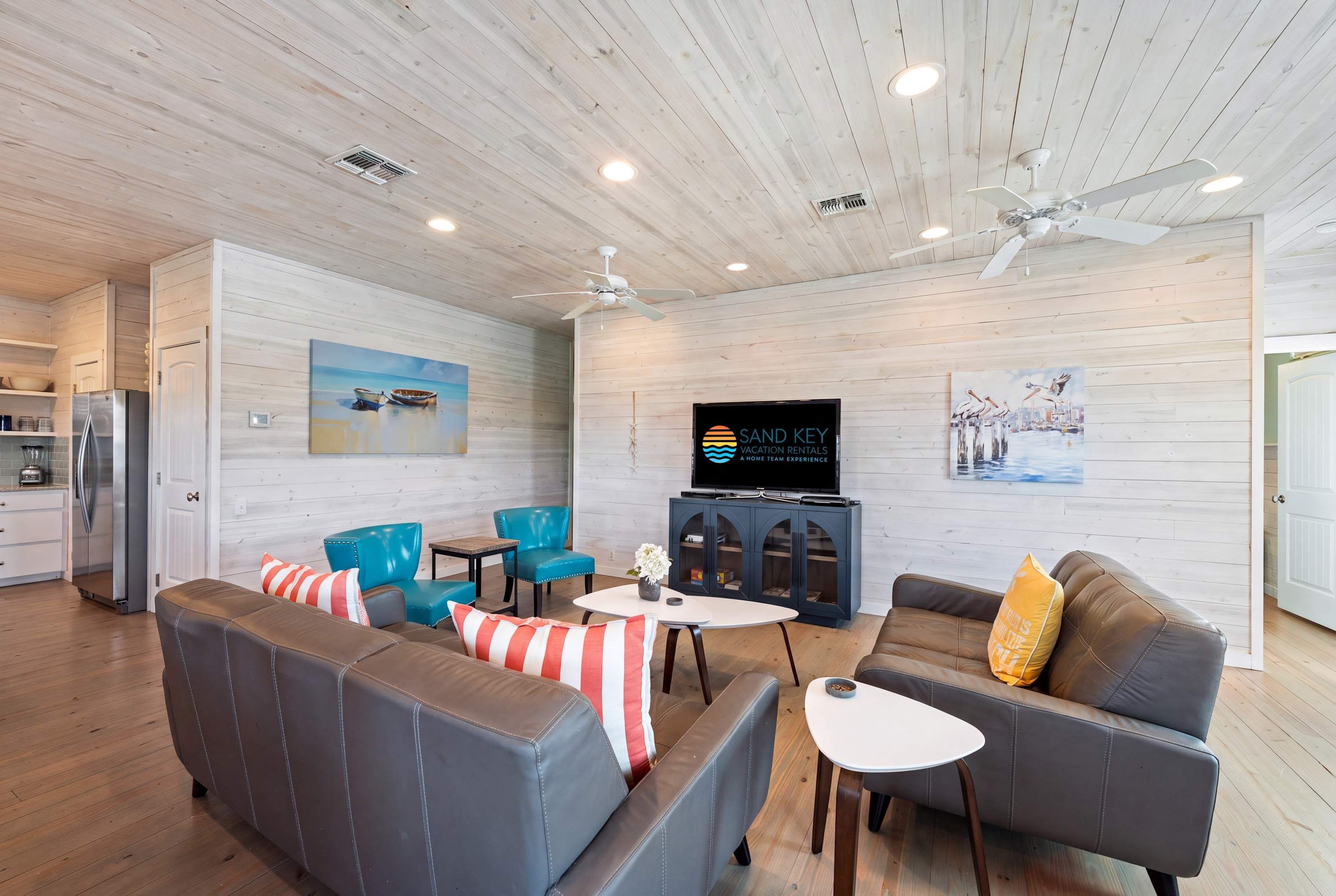A modern living room with a flat-screen TV, leather sofas, a dining table, and a kitchen area with a refrigerator.