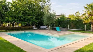 A rectangular swimming pool with a tiled deck, surrounded by grass and trees.