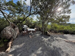 An outdoor dining area with a grill, table, and chairs, surrounded by trees and rocks.