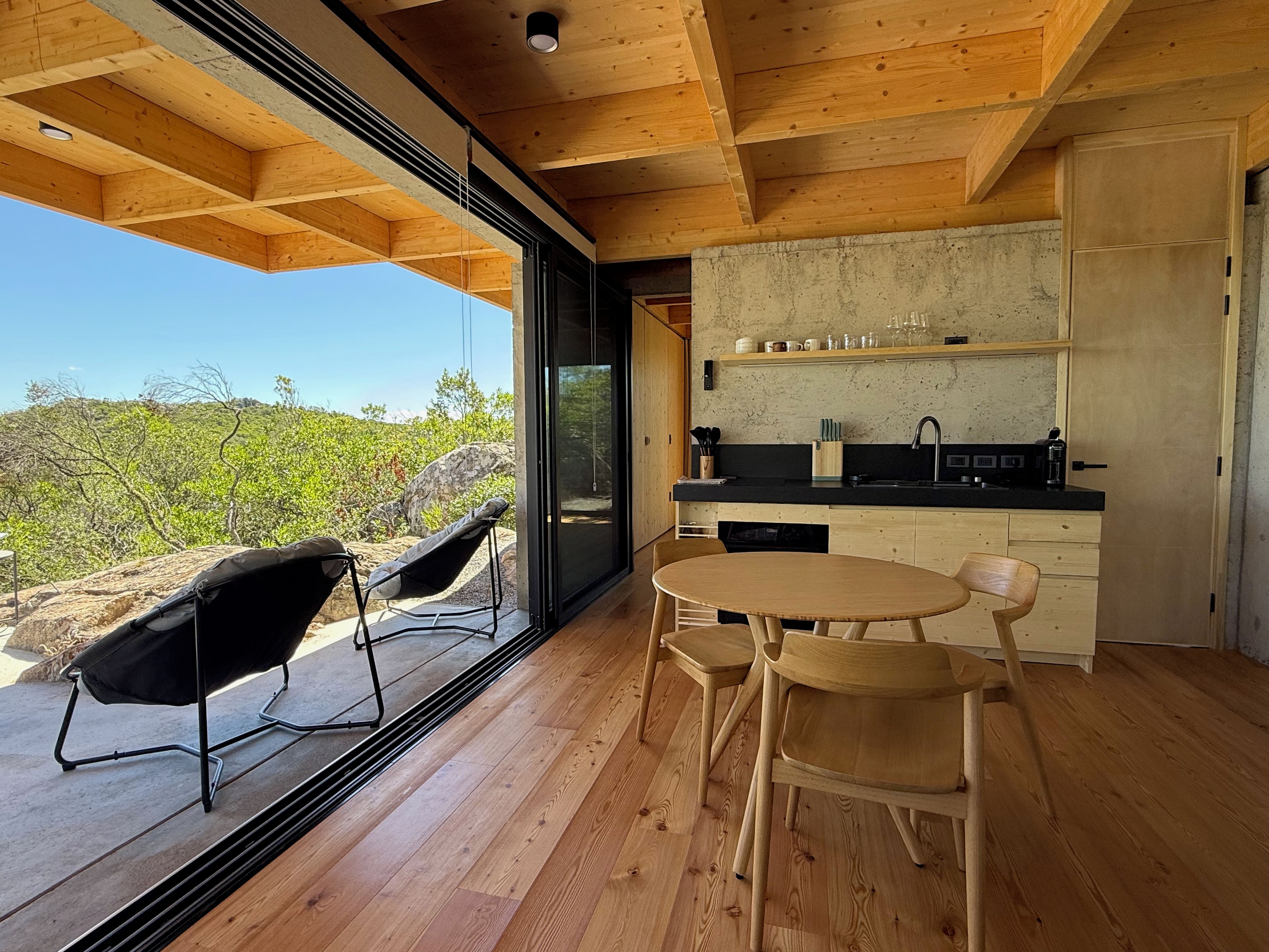 A modern kitchen with a wooden ceiling, a dining area with two chairs, and a large window overlooking a natural landscape.