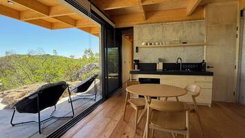 A modern kitchen with a wooden ceiling, a dining area with two chairs, and a large window overlooking a natural landscape.