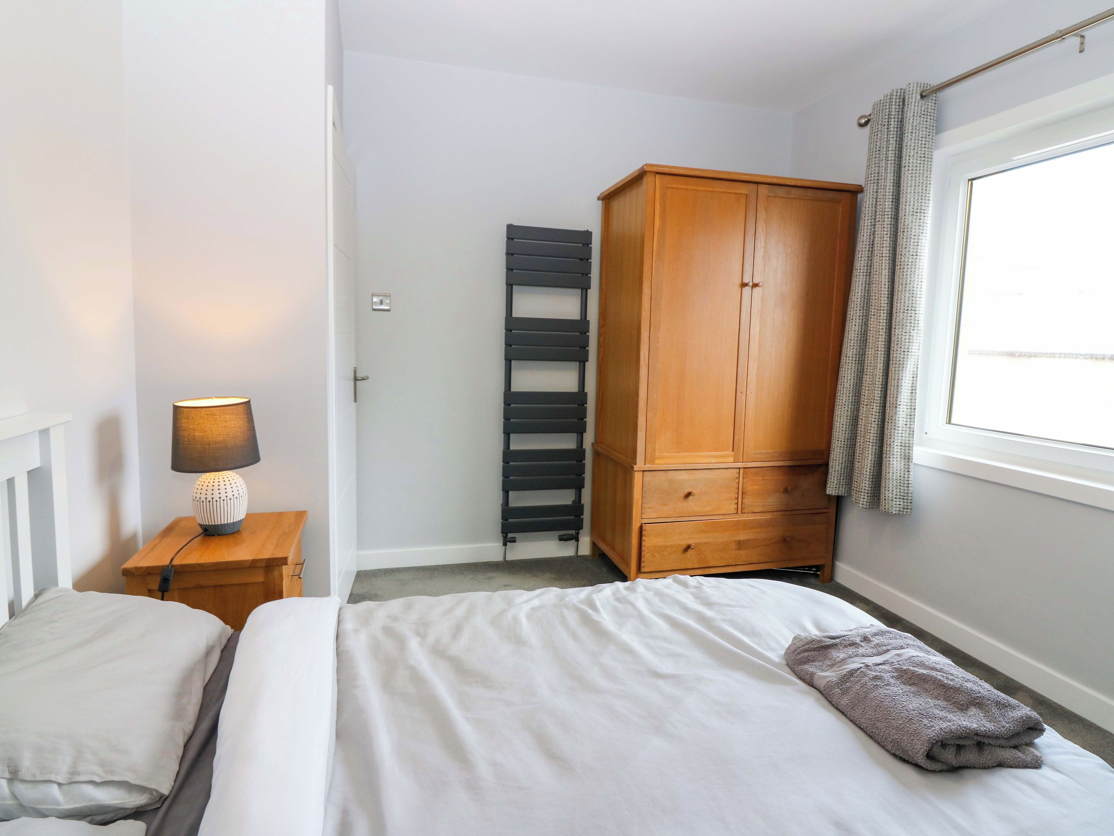 Un dormitorio con una cama, una mesita de noche con lámpara, un armario y una ventana con cortinas.