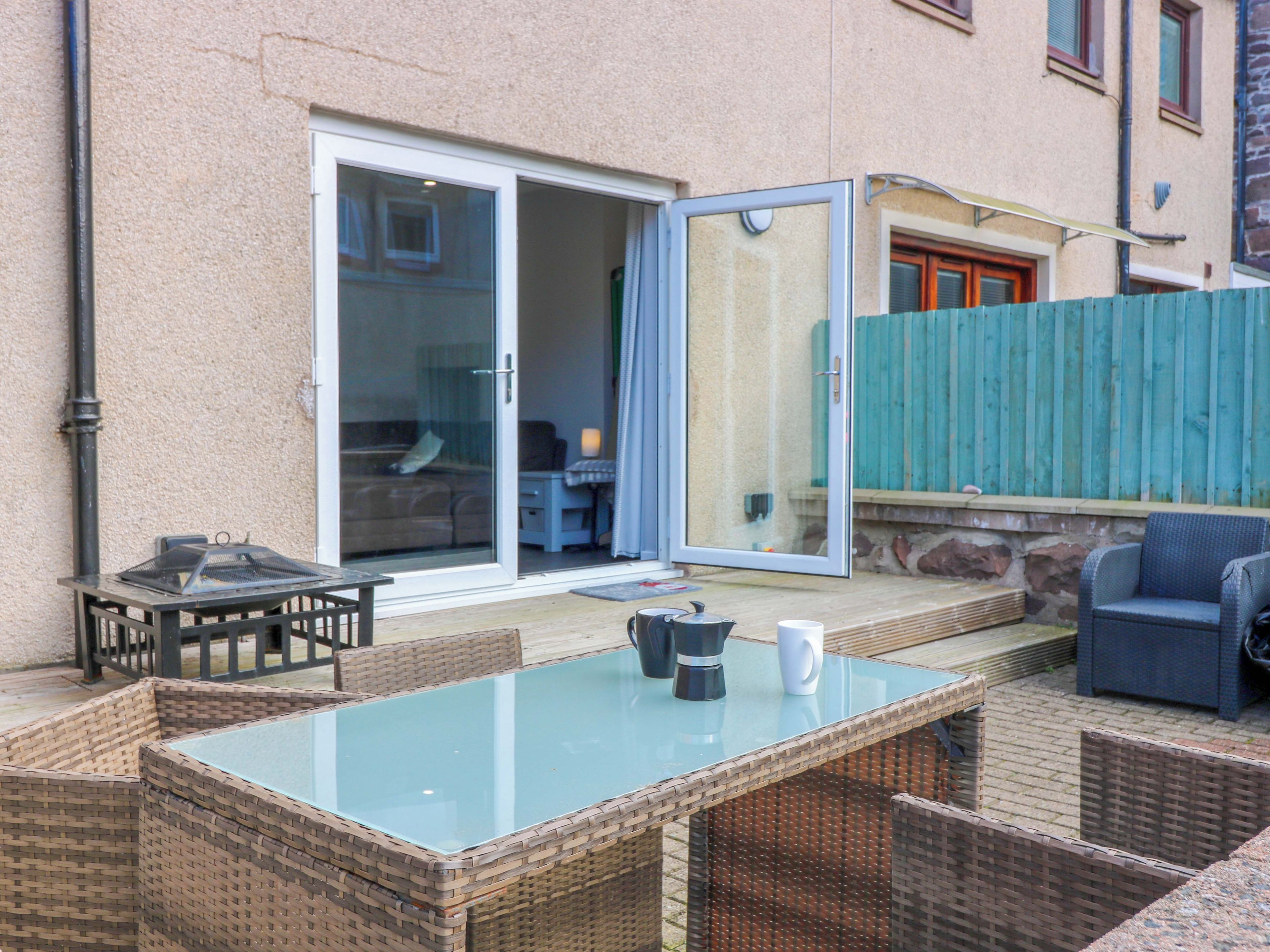 Un patio exterior con una mesa de vidrio, sillas de mimbre, una silla azul y una puerta corrediza de vidrio que da a una habitación.
