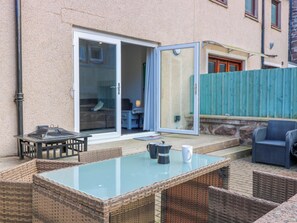 Eine Terrasse im Freien mit einem Tisch mit Glasplatte, Korbstühlen, einem blauen Stuhl und einer Glastür, die zu einem Raum führt.