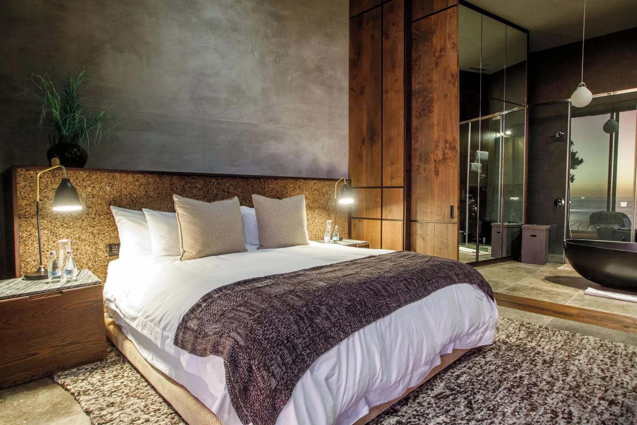 Une chambre moderne avec un grand lit, une table de chevet, une lampe fixée au mur et une vue sur une salle de bain avec une baignoire.