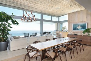 Een ruime woonkamer met een grote houten eettafel, comfortabele zithoek en uitzicht op de oceaan.