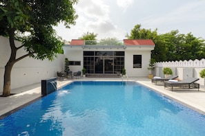A swimming pool with a clear blue water surface, surrounded by a white wall and a small garden area with a tree and a white fence. - ELIVAAS Signature Cabana 3BHK Villa (Jaipur)