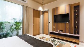 A hotel room with a large bed, a flat-screen TV mounted on a wooden wall, a small desk with a chair, and a large window with curtains.