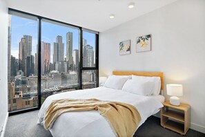 A hotel room with a large window offering a city view, a bed with white linens, a wooden headboard, and two wall-mounted paintings. - Skyline Serenity 2BR in Melbourne CBD, Sky Gym & Pool (Melbourne)