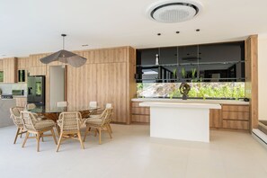 A modern kitchen with a central island, wooden cabinetry, and a dining area with wicker chairs. - New Listing • Villa Bosque del Coco • 4BR Luxury Villa • Coco Bay (Playas del Coco)