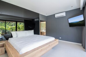 Un dormitorio moderno con una cama grande, mesitas de noche integradas, un televisor de pantalla plana y vistas a un paisaje exuberante.