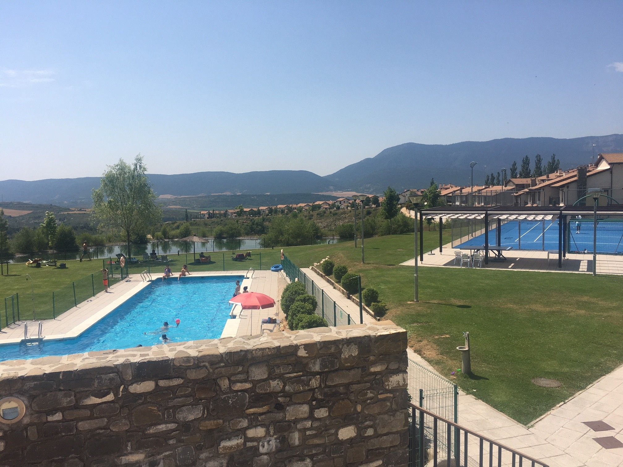 A swimming pool with a stone wall, a red umbrella, and a view of a residential area with houses and mountains in the background.