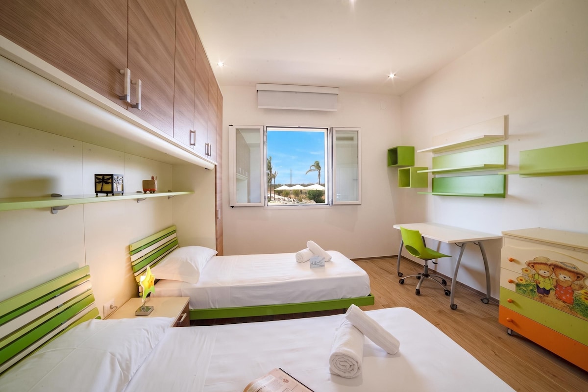 Ein Hotelzimmer mit zwei Betten, einem Schreibtisch, einem Stuhl und einem Fenster mit Blick auf Palmen.