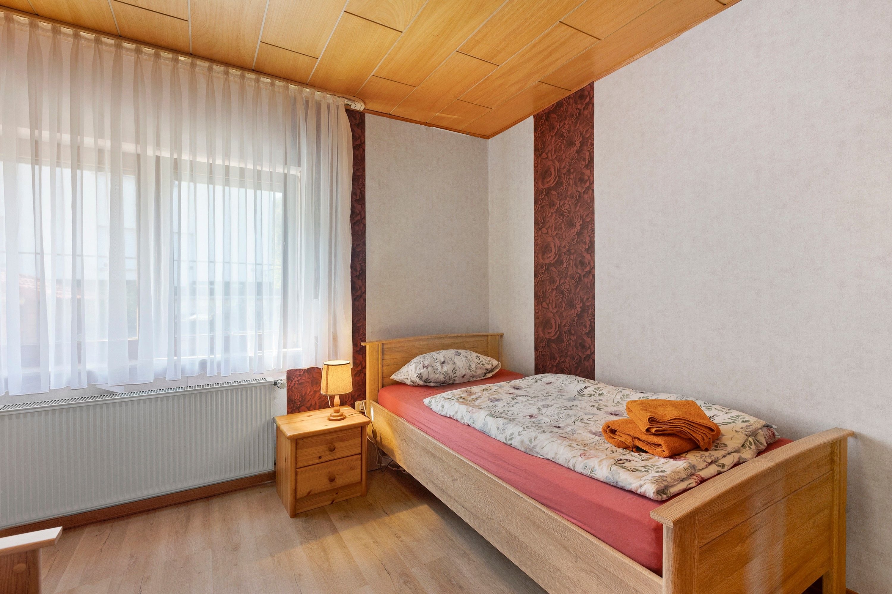 Ein Schlafzimmer mit einem hölzernen Bett, einem Nachttisch mit Lampe, einem Fenster mit durchsichtigen Vorhängen und einer Heizung.
