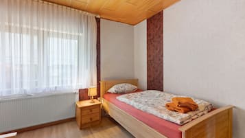 Ein Schlafzimmer mit einem hölzernen Bett, einem Nachttisch mit Lampe, einem Fenster mit durchsichtigen Vorhängen und einer Heizung.