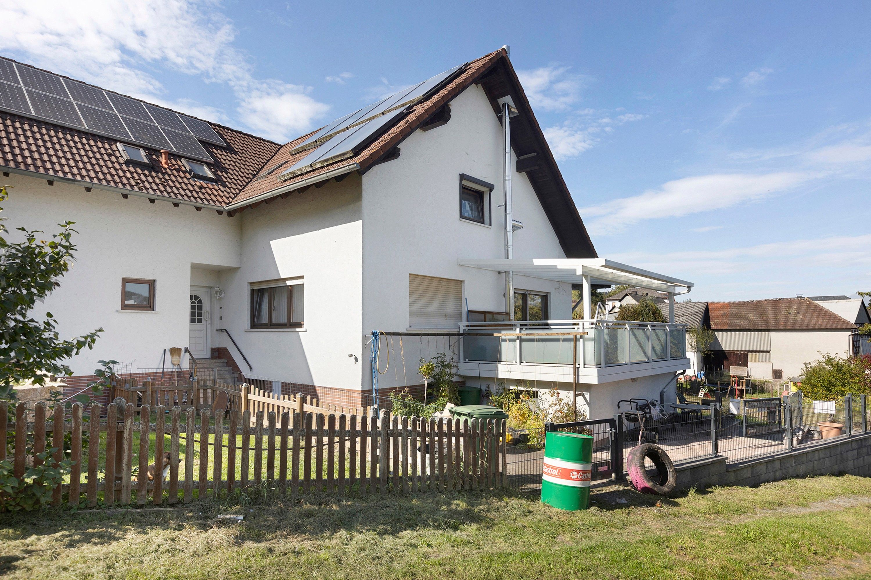 A residential building with a sloped roof, a fenced yard, and a green barrel.