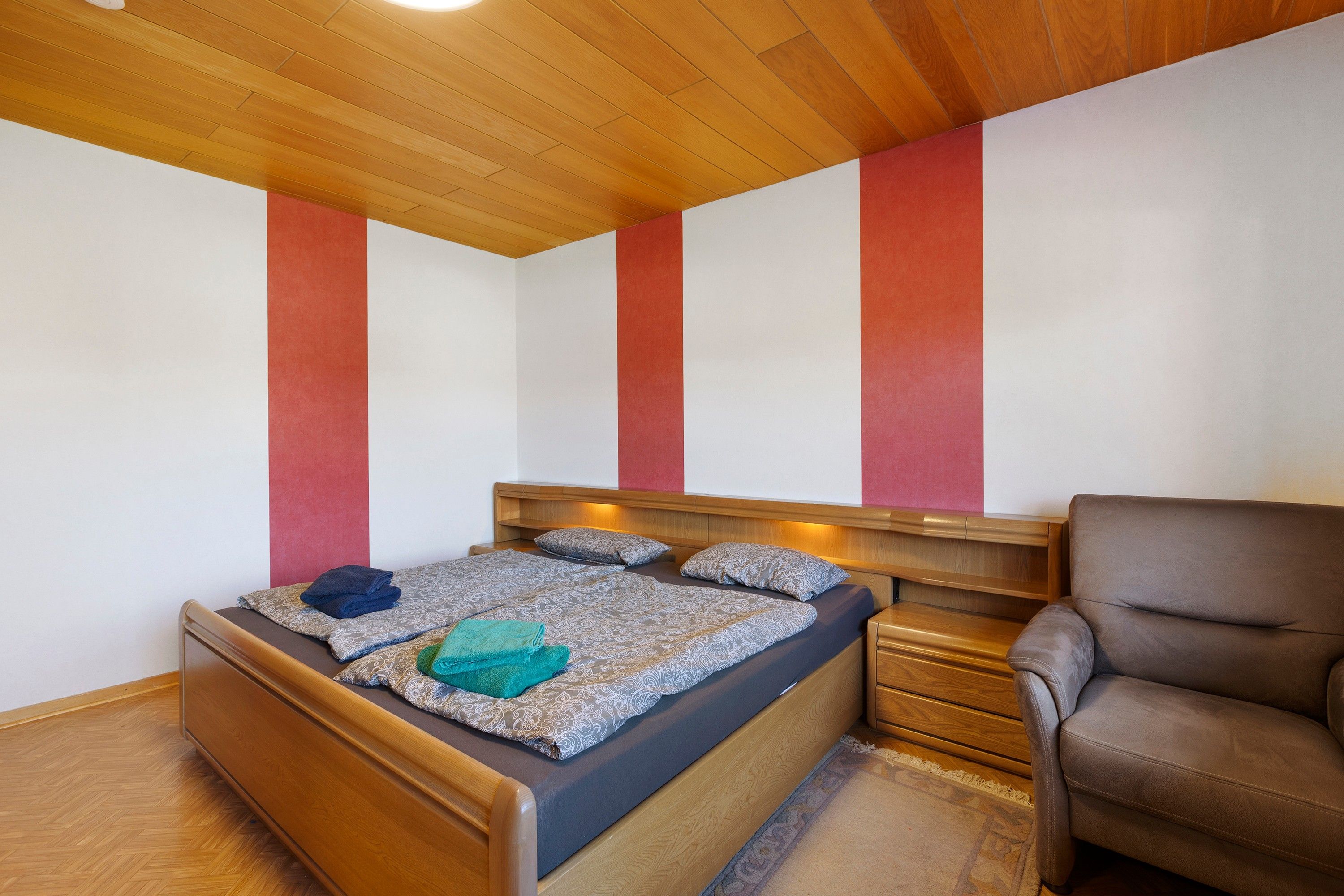 Un dormitorio con cabecera de madera, cama con ropa de cama estampada, un sillón de cuero marrón y suelo de madera.